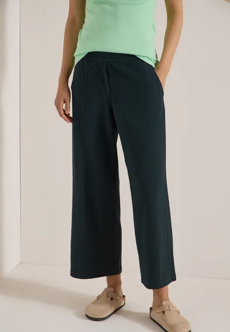 Wijde, geplooide zwarte broek met zakken, groen topje en beige slippers.