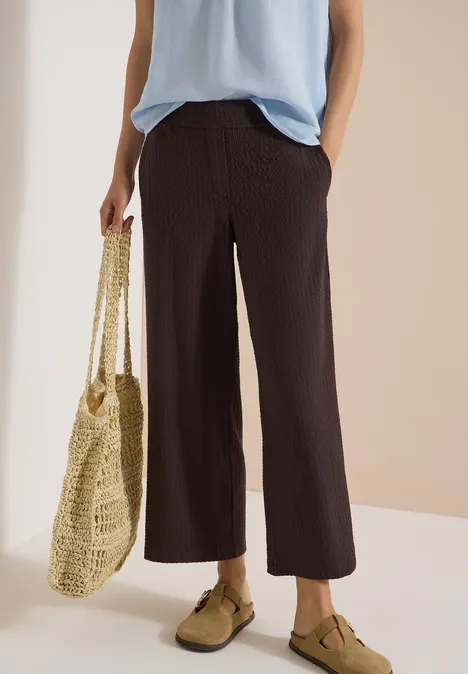 Dame met lichtblauw hemdje, brede bruine ribbroek, groene loafers en gehaakte tas.