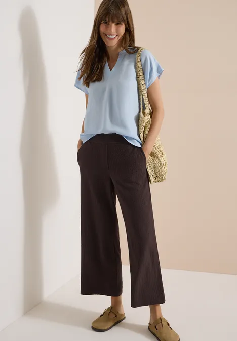 Vrouw in lichtblauw T-shirt, zwarte wijde broek, strozak en bruine loafers voor neutrale achtergrond.