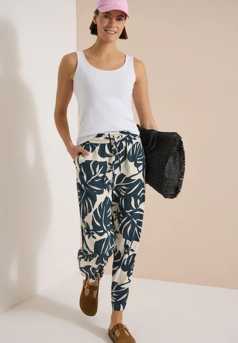 Broek met High Waist en Wide Leg pijpen in een Loose Fit pasvorm palm breeze green Vrolijke vrouw in wit topje, gebloemde broek en roze pet, met een zwarte strandtas.