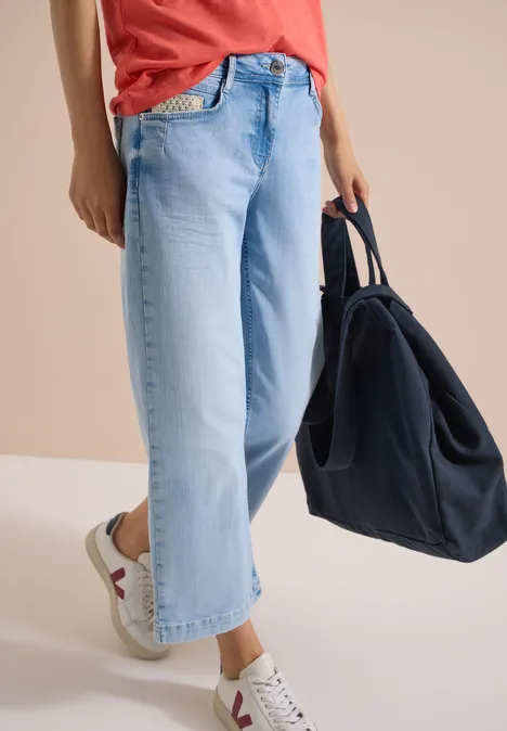 Jean 7/8 Wide Leg avec détail crocheté light blue random bleached Person von der Taille abwärts in hellblauen, weiten Jeans mit weißen Sneakern und dunkelblauer Tasche.