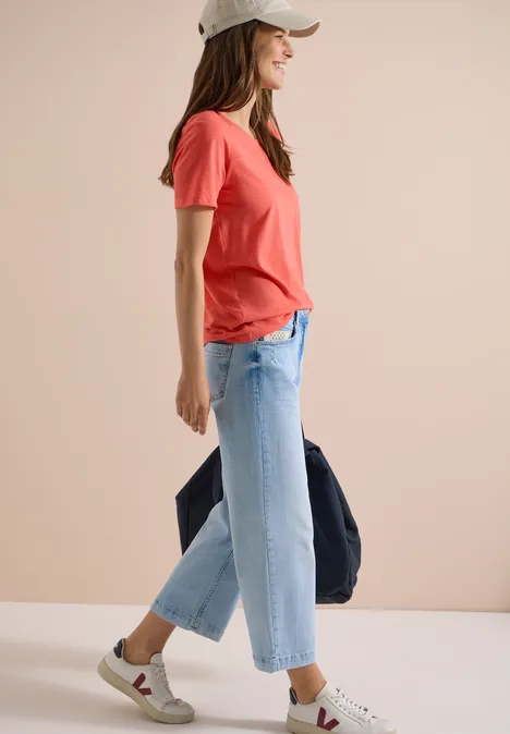 Jean 7/8 Wide Leg avec détail crocheté light blue random bleached Frau im Profil mit rotem T-Shirt, hellblauen Jeans, weißen Sneakers und Cap, hält eine dunkle Jacke.