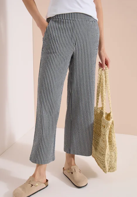 Person in weißem Top, gestreifter, weiter 7/8-Hose, beigen Slippern und geflochtener Basttasche.