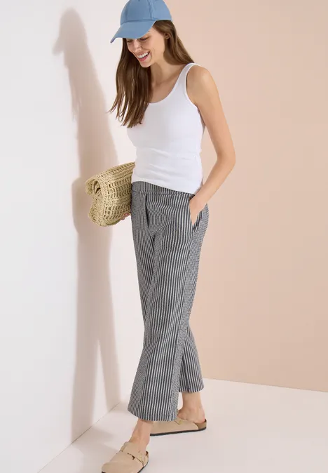 Frau in weißem Top, grauen, karrierten Hosen, blauem Cap und Strohtasche lehnt lässig an der Wand.