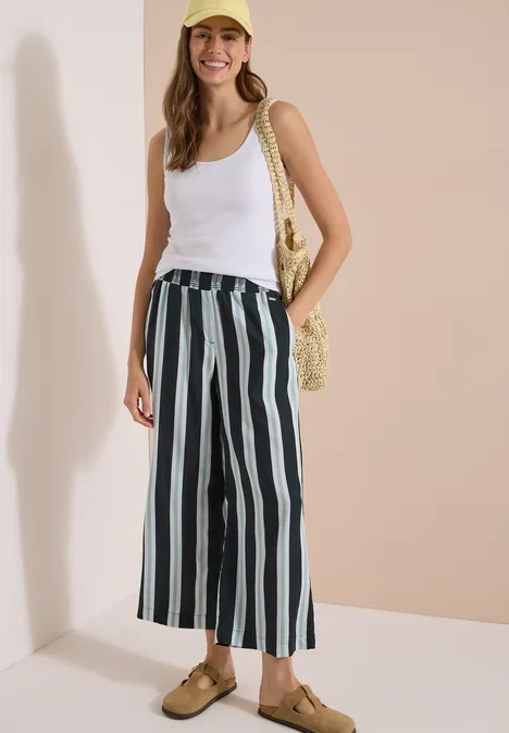 High Waist Wide Leg Hose mit Streifen palm breeze green Frau in weißem Tanktop, gestreifter Marlenehose, Strohtasche und Sandalen vor hellem Hintergrund.