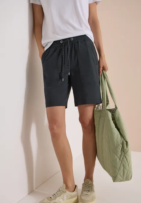 Person in weißem T-Shirt, schwarzen Shorts und Sneakers mit großer, gesteppter grüner Tragetasche.