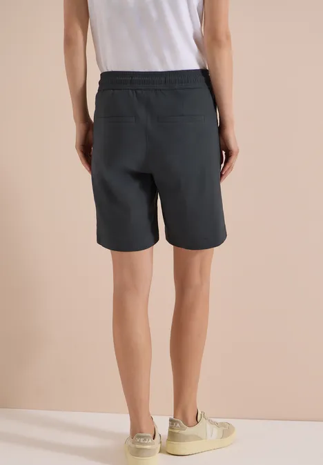 Rückansicht einer Person in weißem Top, schwarzen Shorts und beigen Sneakers vor hellem Hintergrund.