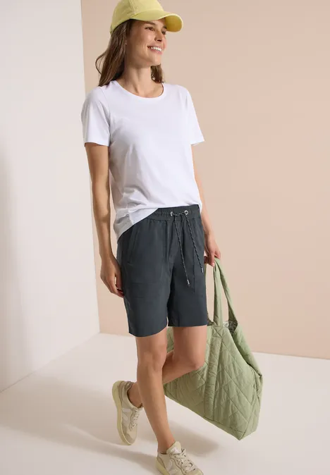 Frau in weißem T-Shirt, grauen Shorts und beigen Sandalen hält eine grüne Tasche.