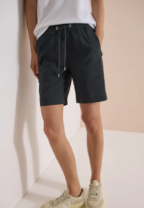 Frau in schwarzen, knielangen Shorts mit Kordelzug, weißem Shirt und hellen Sneakern vor minimalistischem Hintergrund.