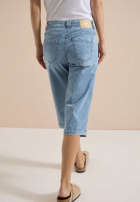 Personne de dos porte un pantalon en jean bleu clair coupé aux genoux et des sandales sur fond beige.