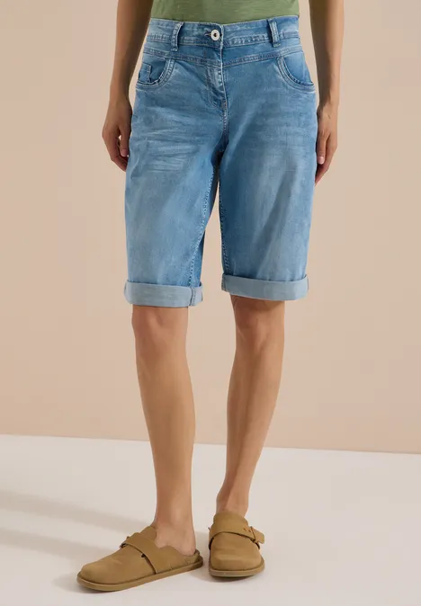 Une personne porte un bermuda en jean bleu clair et des sandales beiges devant un fond neutre.