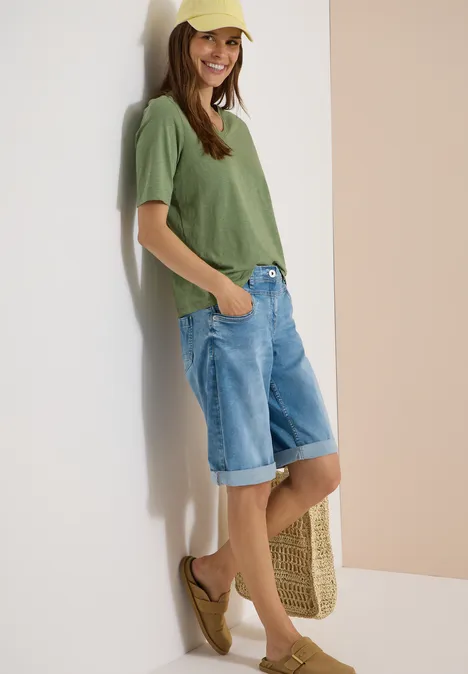 Jeune femme appuyée contre un mur, vêtue d'un t-shirt vert et d'un short en jean au genou.