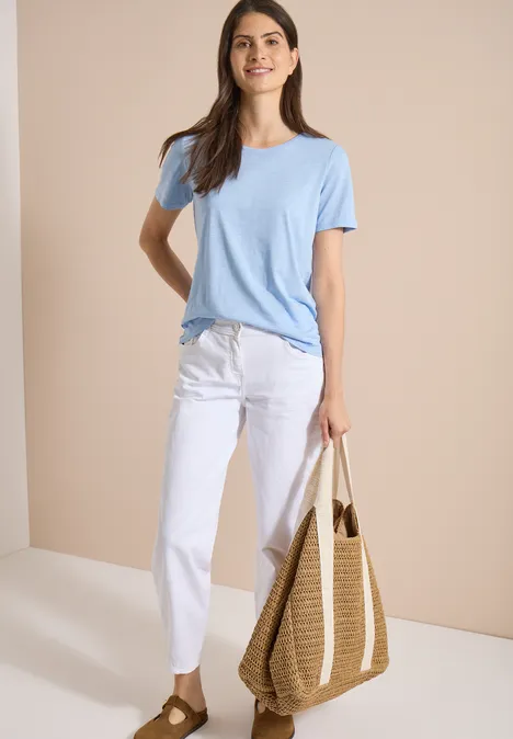 Jean taille haute à Barrel Leg, coupe ample White Femme en t-shirt bleu clair, pantalon blanc, tenant un sac en osier sur fond beige.