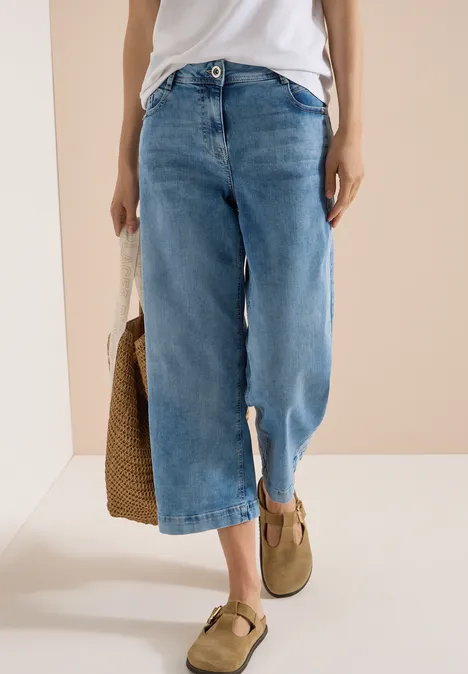 Vrouw in wit topje met wijde denim culotte met split, beige loafers en een gevlochten rieten tas.