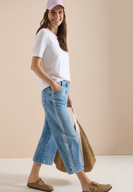 Vrouw in wit T-shirt, wijde korte jeans, rieten tas en platte sandalen voor neutrale achtergrond.