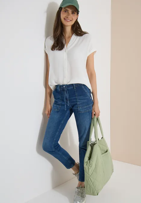 Straight Leg Jeans mit Gürteldetail mid blue used wash Frau in weißem T-Shirt, blauen Jeans und Baseballcap mit grünem Shopper, die entspannt an einer Wand lehnt.