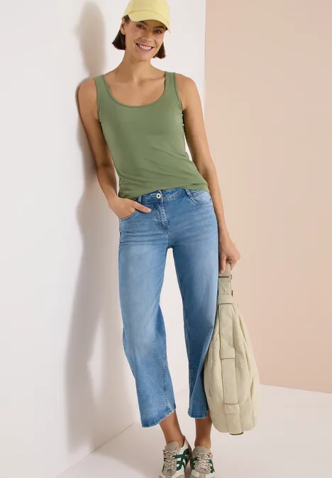 Een persoon met een groen topje en blauwe jeans leunt tegen een muur en houdt een lichte tas vast.