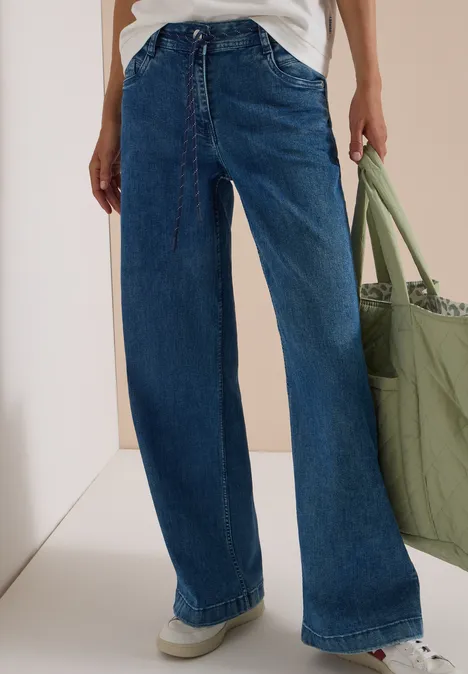 Jean bleu ample taille haute, jambes larges, associé à un top blanc et un grand sac vert en tissu.