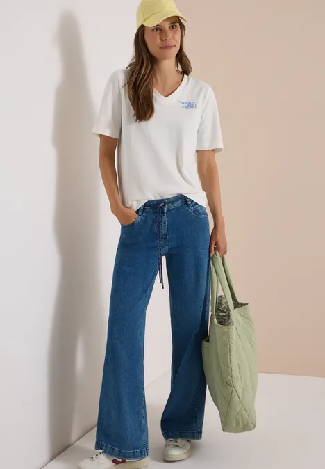 Une femme porte un T-shirt blanc à col en V, un jean flare bleu, une casquette jaune pâle et tient un sac vert olive.