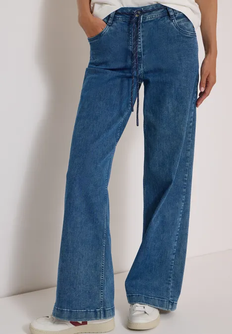 Personne portant un jean bleu taille haute, coupe évasée et baskets blanches.