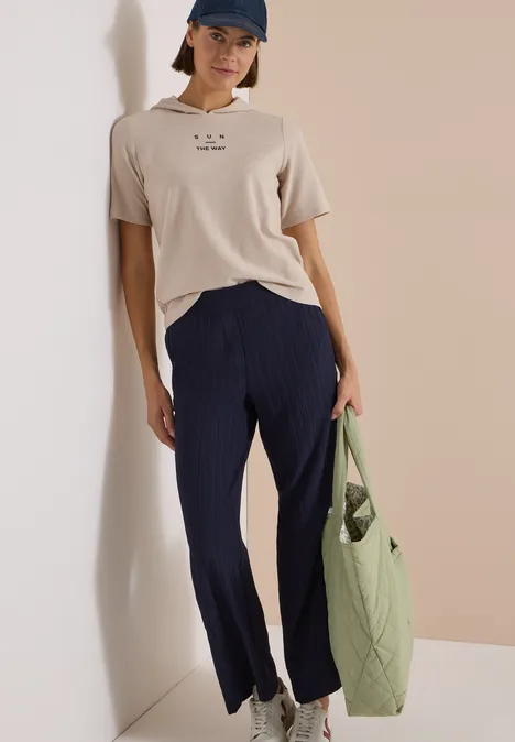 Wide Leg Hose mit Strukturstreifen urban dark blue Frau trägt beige T-Shirt, dunkelblaue Hose, blaue Kappe und hält olivgrüne Stepp-Tasche.