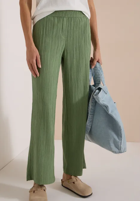 Ein Modell trägt grüne Plissee-Hose mit weitem Bein und hoher Taille, dazu weiße Schuhe und Jeansjacke.