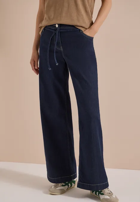 Frau trägt weite, dunkelblaue Jeans mit Kordelzug und Sneakers vor hellem Hintergrund.