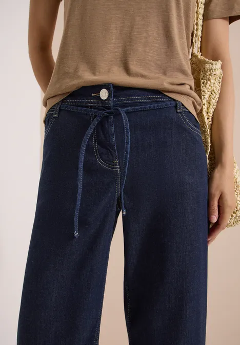 Person trägt hoch geschnittene, dunkelblaue Jeans mit Bindegürtel, kombiniert mit braunem Oberteil und Basttasche.