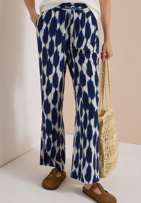 Pantalon ample bleu-blanc à motif ikat, haut blanc, sac en osier et mocassins marron.