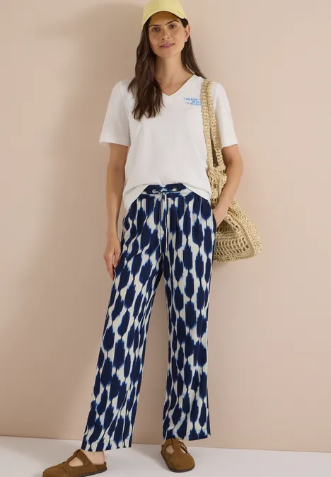Femme en t-shirt blanc col V, pantalon ample bleu à motifs blancs et sac en osier.