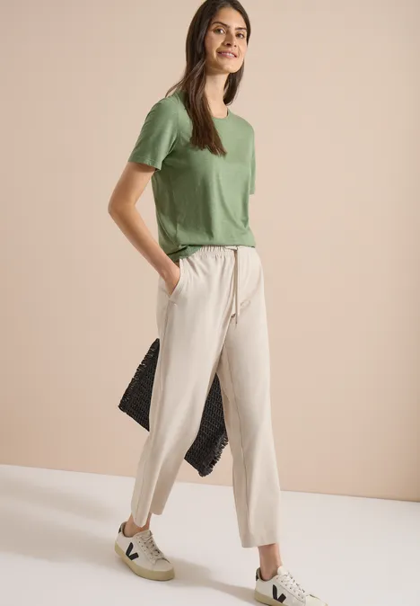 Femme portant T-shirt vert, pantalon beige et baskets blanches, posant de profil devant un mur beige.