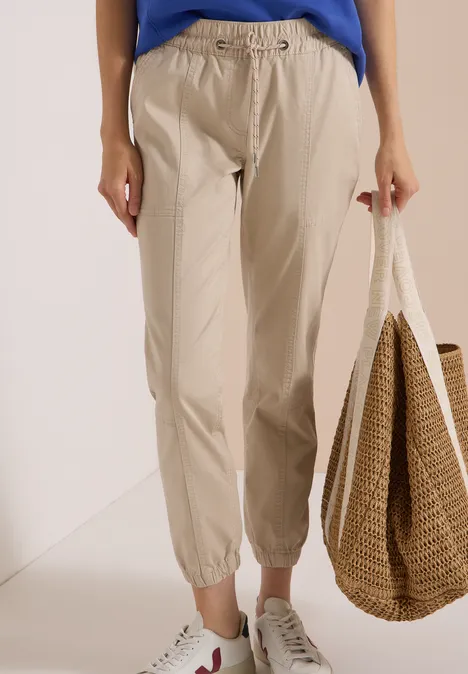 Frau trägt helle, beige Hose mit Gummizug, weiße Sneaker und hält eine geflochtene Tasche.
