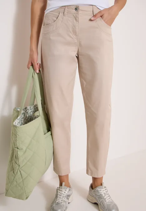 Barrel Leg Hose in Papertouch Qualität cashmere beige Eine Person trägt beige Chinohose, weiße Sneaker und hält eine grüne, gesteppte Tasche.