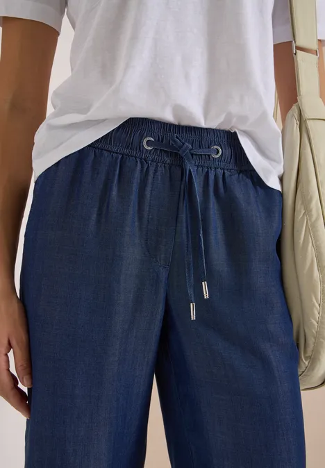 Person trägt helles T-Shirt, dunkelblaue, lockere Hosen mit Kordelzug und eine helle Schultertasche.