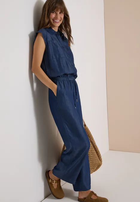 Eine Frau lehnt an einer Wand, trägt einen blauen Jeans-Jumpsuit, Basttasche und braune Loafers.