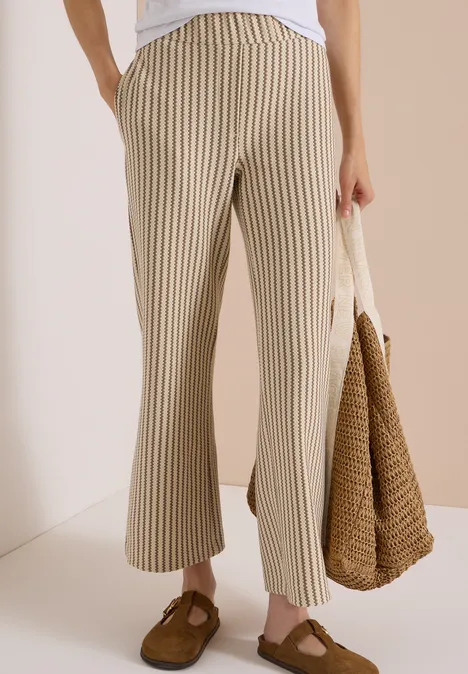 Beine in beigen, gestreiften Hosen, braune Loafers und eine Strohtasche in der Hand.