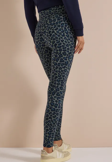 Eine Person trägt blaue Leggings mit dunkelblauem Leopardenmuster und hellen Sneakern vor beigem Hintergrund.