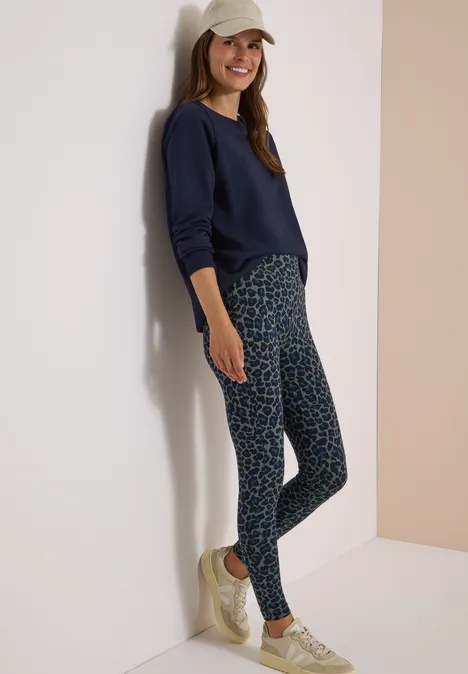 Frau mit Mütze steht an Wand, trägt dunkelblaues Sweatshirt, gemusterte Leggings und helle Sneaker.