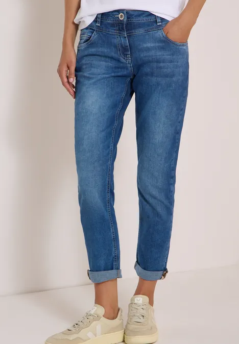 Frau trägt blaue Jeans mit umgeschlagenen Säumen und weiße Sneakers vor neutralem Hintergrund.