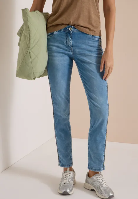 Frau in hellblauen Jeans, braunem Top und grauen Sneakern trägt eine grüne Steppjacke über dem Arm.