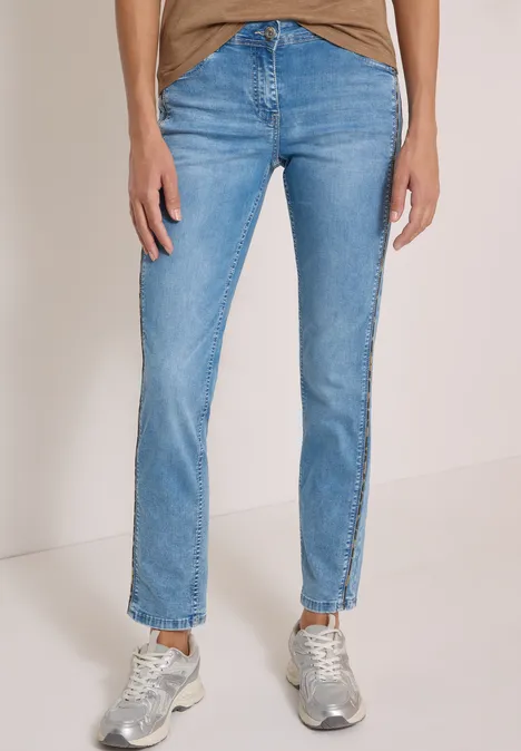 Person in hellblauen Jeans und silbernen Sneakers vor einfarbigem Hintergrund.