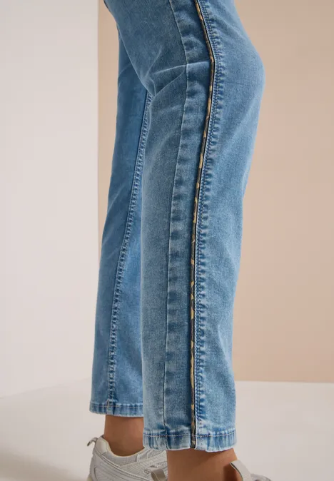 Schlanke blaue Jeans mit seitlichem Saumschlitz, kombiniert mit beigefarbenen Sandalen vor neutralem Hintergrund.