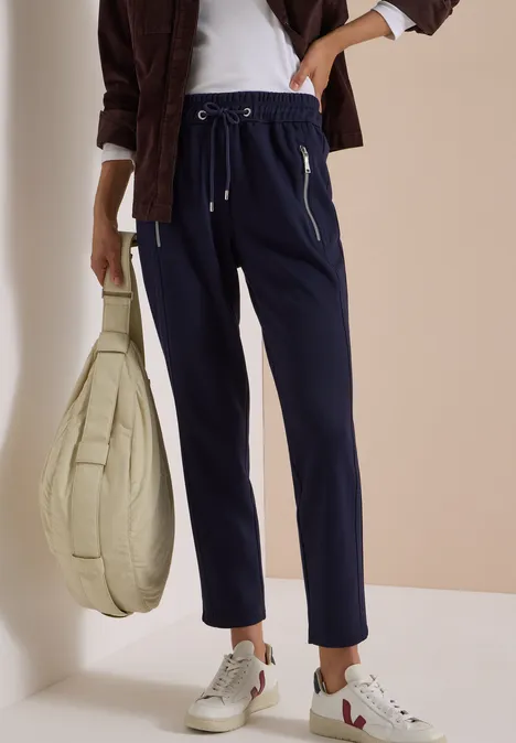 Personne en joggings bleu marine avec poches zippées, baskets blanches et grand sac beige matelassé.