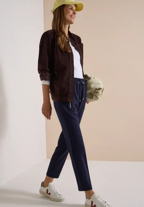 Une femme en casquette verte, veste marron, t-shirt blanc, pantalon bleu et baskets blanches tient un sac.
