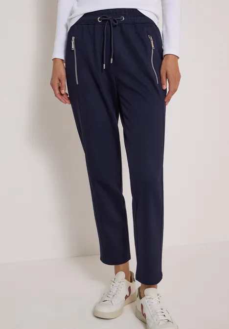 Pantalon femme bleu marine à taille élastique, cordon et poches zippées dorées, avec baskets blanches.