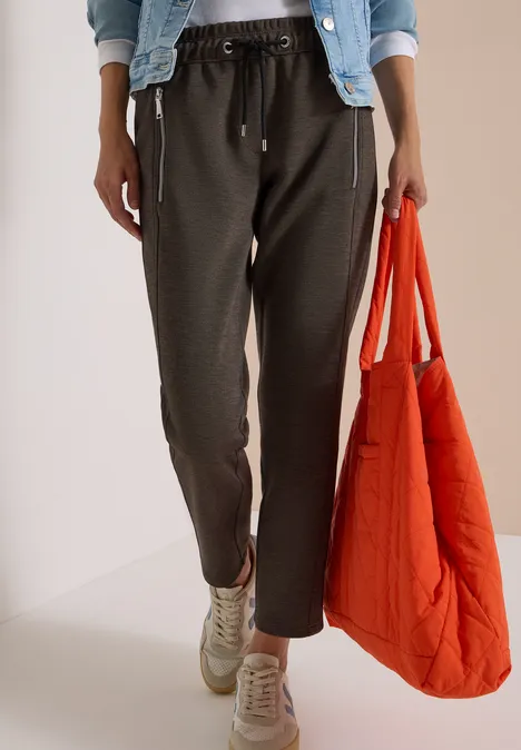 Personne porte veste en jean, haut blanc, pantalon jogging noir, baskets et porte un sac matelassé orange.