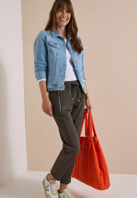 Femme souriante en T-shirt blanc, veste en jean, pantalon de jogging gris foncé, baskets blanches et sac rouge.