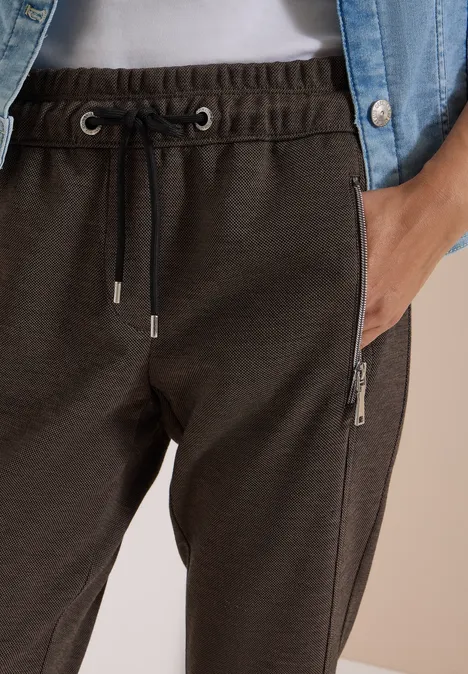 Jogging foncé à taille ajustable avec poches zippées latérales, porté sous une chemise en jean.