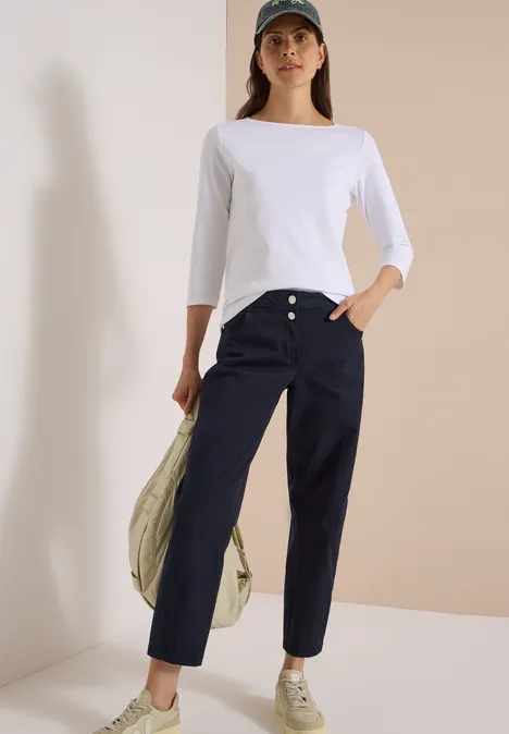 Femme en t-shirt blanc, pantalon bleu marine, baskets et casquette, tenant une veste sur l’épaule.