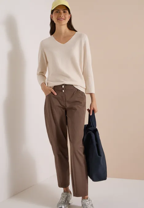 Mid Waist Barrel Leg Hose im Casual Fit urban taupe Een vrouw draagt een licht V-hals trui, bruine broek en pet, met een marineblauwe tas.